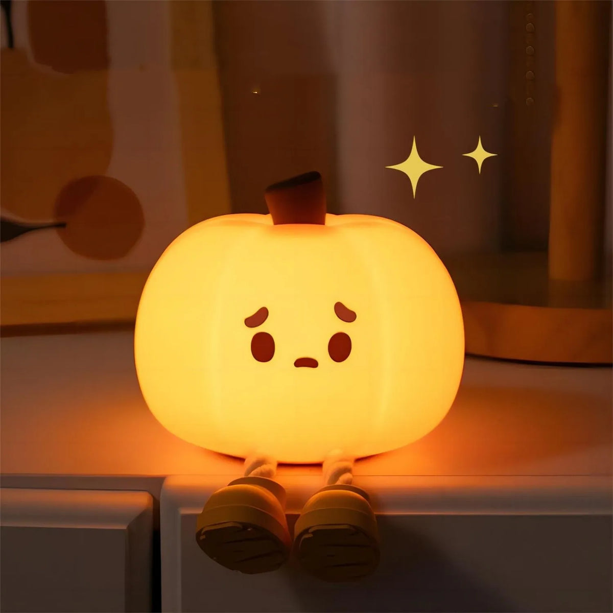 Halloween Pumpkin Silicone Night Light — glowing pumpkin with cute sad face sitting on a bedside table with legs dangling, warm orange glow in a bedroom