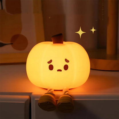 Halloween Pumpkin Silicone Night Light — glowing pumpkin with cute sad face sitting on a bedside table with legs dangling, warm orange glow in a bedroom